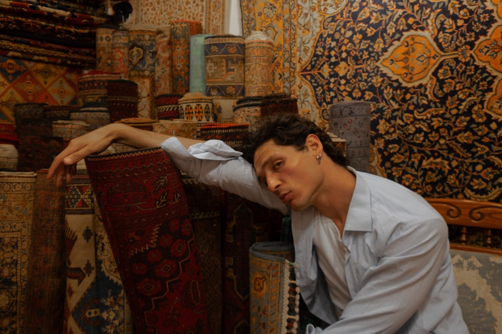 An artisan in an open linen shirt leans against towering rolled rugs in the Professio archive